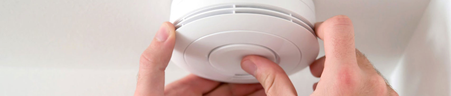 A person's hand reaches up to press the test button on a smoke alarm.
