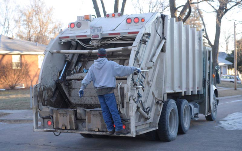 A garbage truck on a street with a person riding on the back.