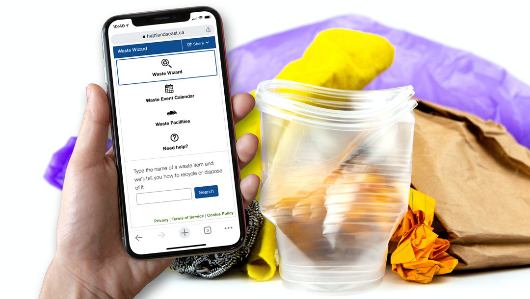 Hand holding an iPhone with the Highlands East Waste Wizard pulled up on it. Some garbage is in the background.